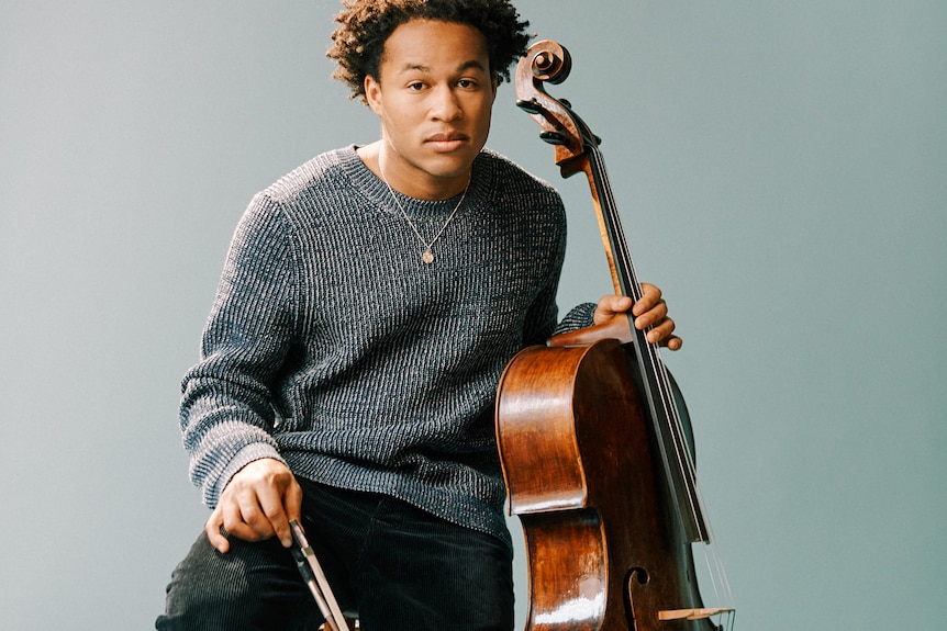 A young man sits on a stool, wearing a grey jumper and holding his cello to one side.