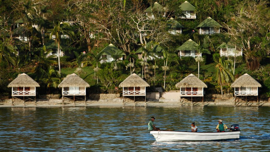 Three people commute in a small metal boat on calm ocean in front of villas that sit on a green mountainside close to the ocean