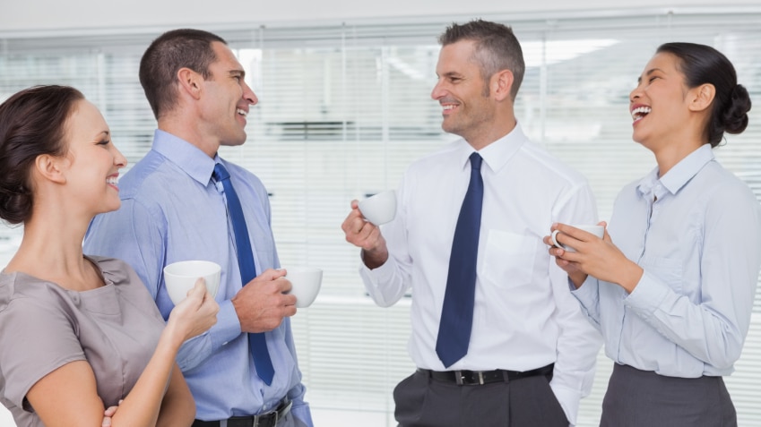 Office workers standing around and joking while holdings cups of coffee.