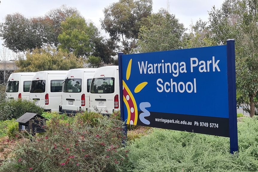 A school sign and a row of four white mini-buses. 