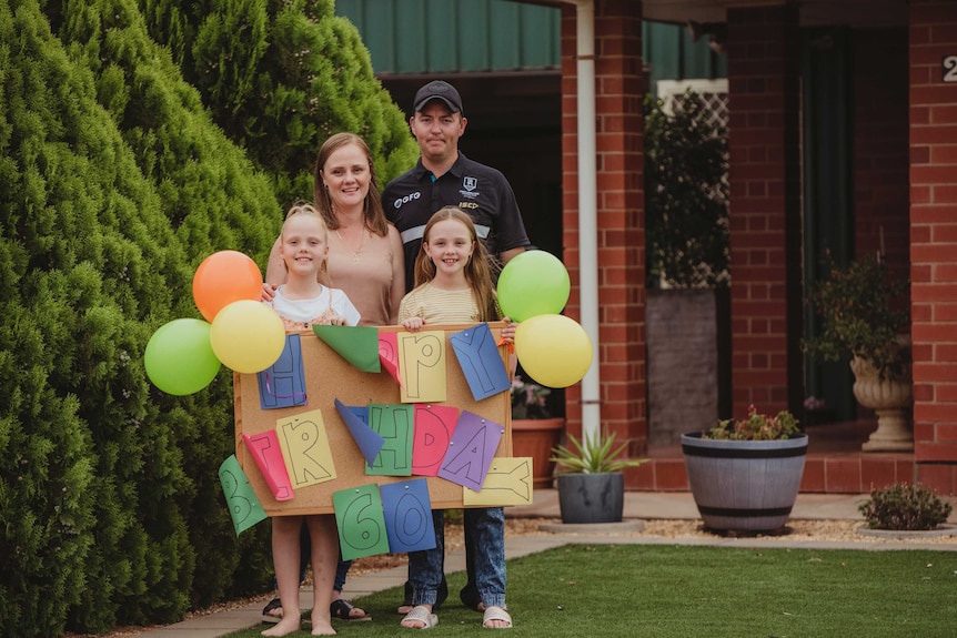 A Whyalla family stands outside their house with a handmade sign that reads 'Happy Birthday 60', surrounded by balloons.