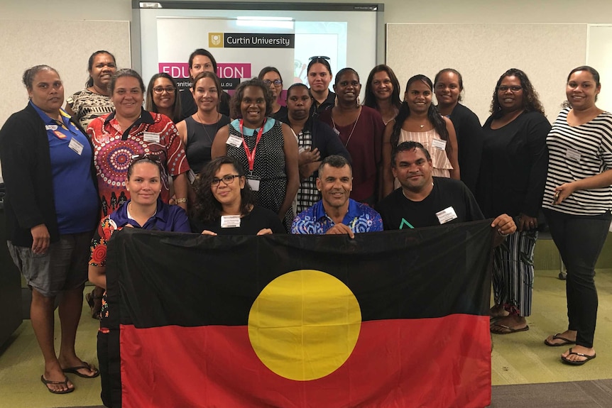A group of Indigenous people in a classroom holding up the Aboriginal flag.