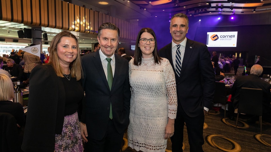 Smiling two men, wearing suits and ties, and two women wearing dresses inside a convention hall.