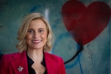 A woman wearing a red blazer smiles, standing in front of an art piece with a red heart.