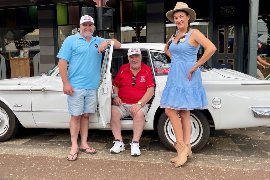 Two men and a woman, all smiling as they pose around a vintage car.