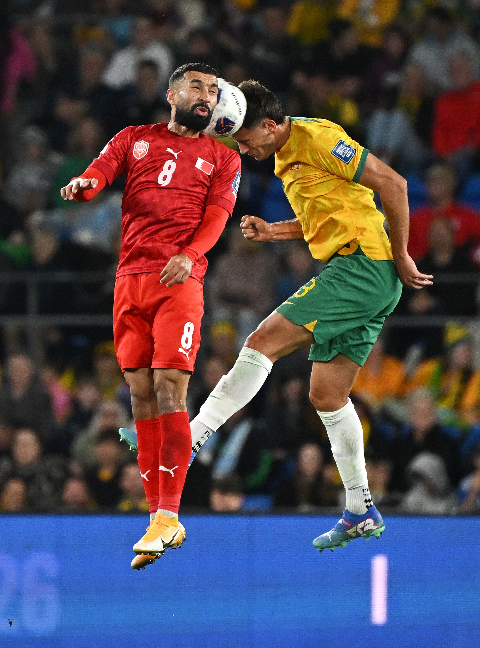 One man wearing a red jersey and one man wearing a green and gold jersey collide head-first with a ball between them.