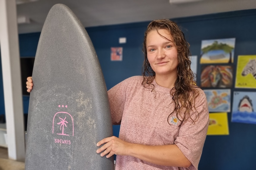A young woman, wearing a pink tee-shirt, holding a gray surfboard.