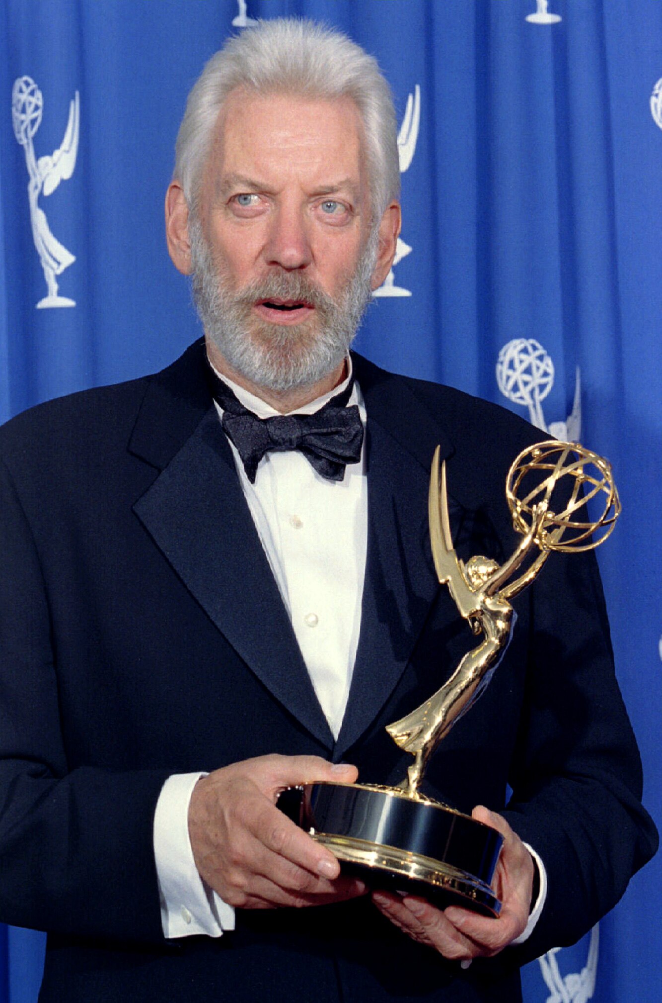 Donald Sutherland in a navy suit standing and holding his Emmy award