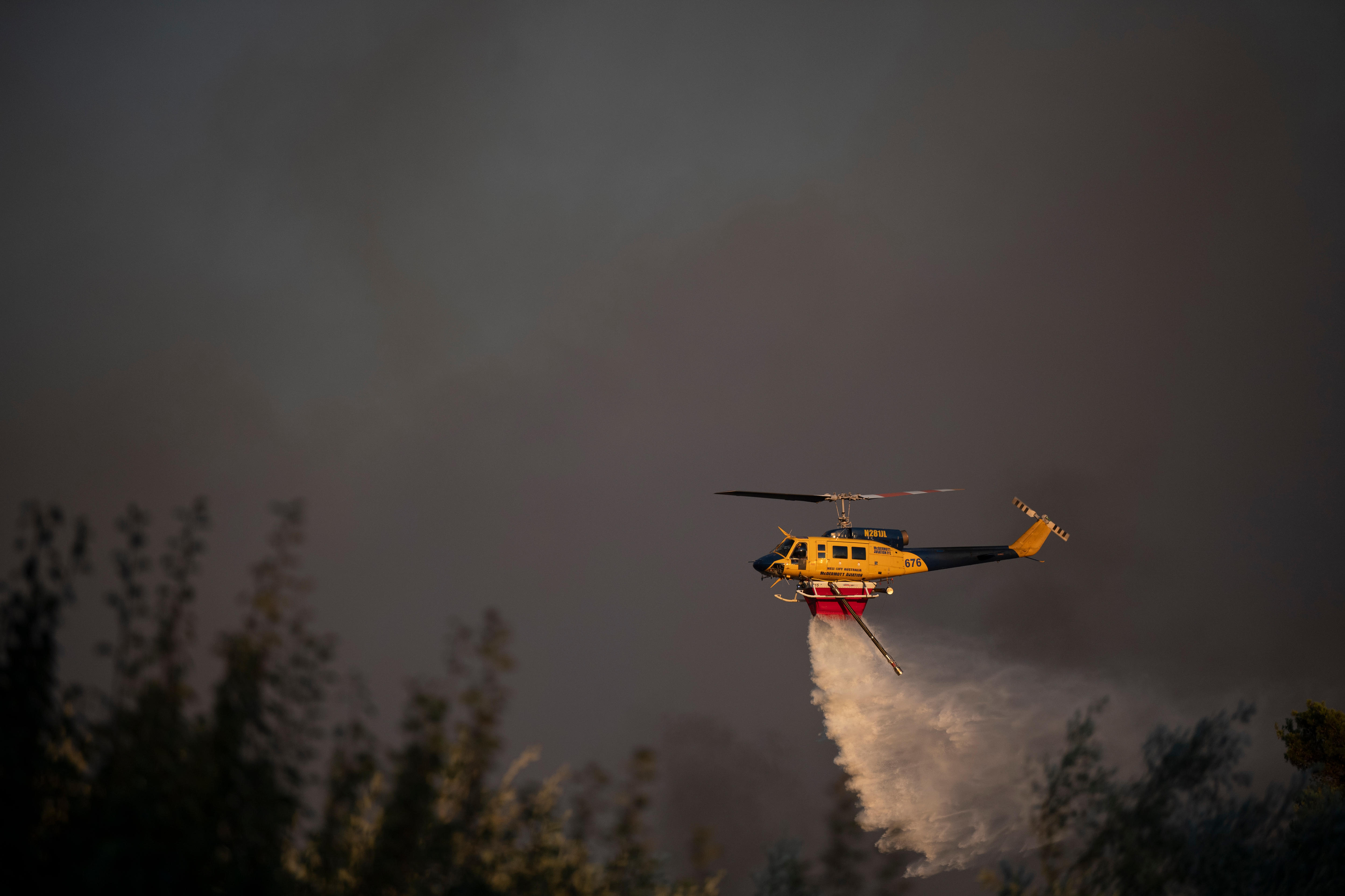 A yellow helicopter drops water on a forest, clouded in smoke.