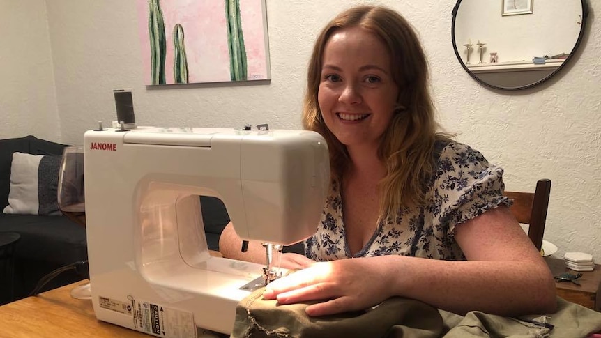 A young woman with strawberry blonde hair smiles as she stitches a linen skirt on her sewing machine, which is on a table