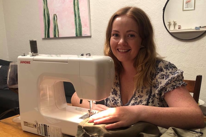 A young woman with strawberry blonde hair smiles as she stitches a linen skirt on her sewing machine, which is on a table