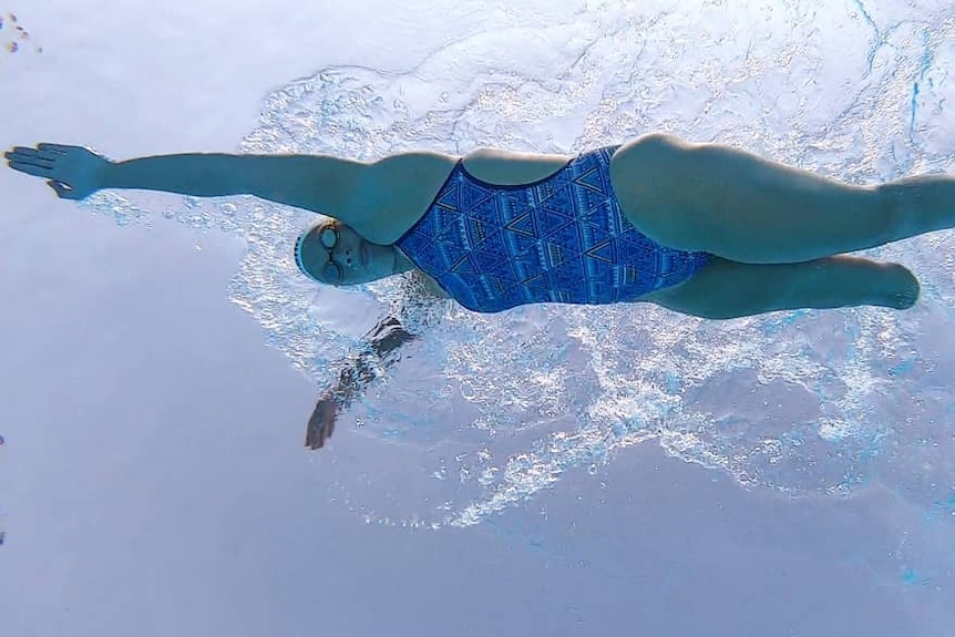 A woman is wearing blue swimmers and doing freestyle in a pool. She is amputated below the right knee.