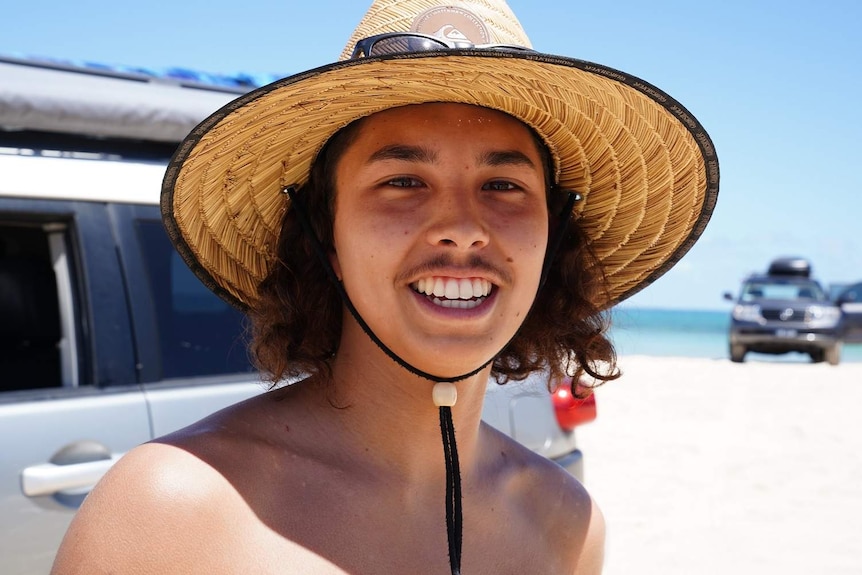 Tyler Hammill smiles wearing a straw hat on a beach with 4WDs in the background.