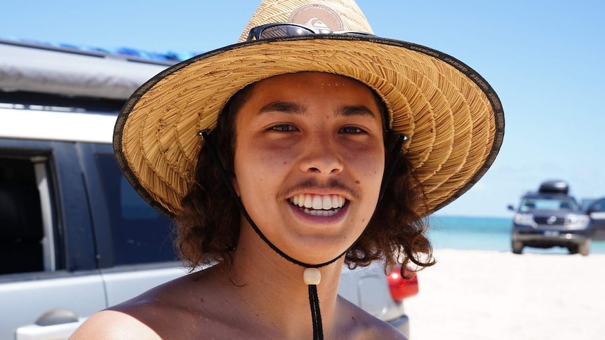 Tyler Hammill smiles wearing a straw hat on a beach with 4WDs in the background.