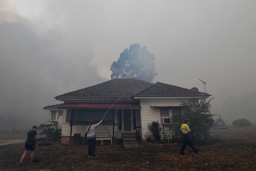 A woman stands outside her single-story home hosing down its roof for embers. Smoke fills the sky.