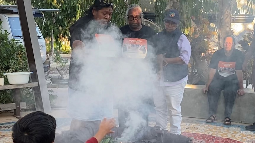 three women stand arm in arm surrounded by smoke from a ceremonial fire, while a child reaches for the smoke