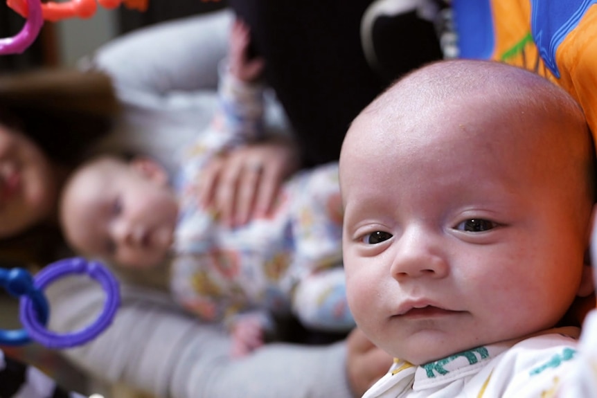 A baby in the foreground of the shot and another baby and their mother in the background