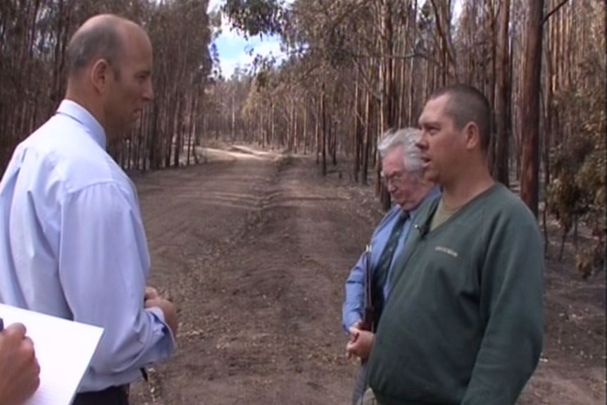 A detective wearing a business shirt speaks with a man in a green jumper while the pair stand in a charred forest