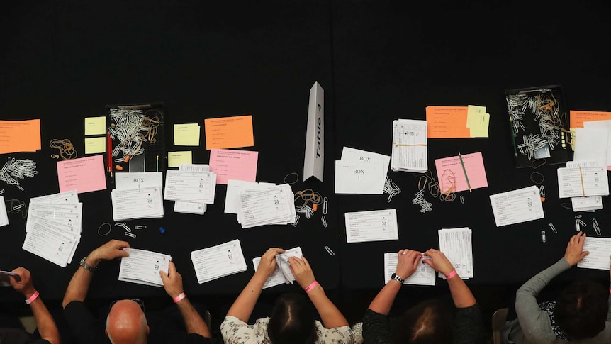 An overhead shot of five people sitting sorting ballot papers which are white, pink and yellow.