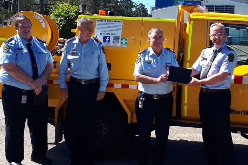 Four men in rural fire brigade uniforms with a tablet, standing in front of a yellow fire truck