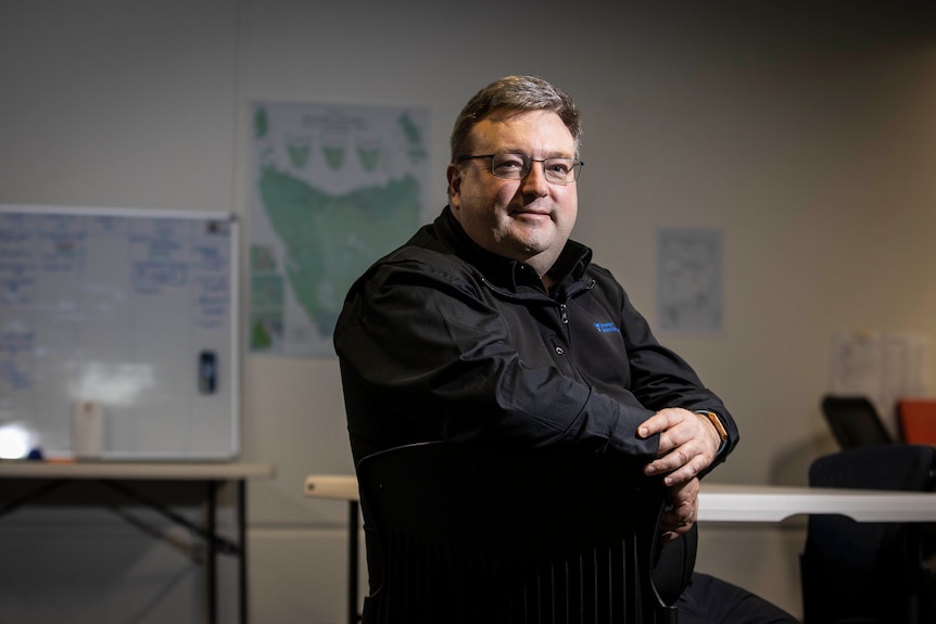 Middle aged man in black shirt and glasses sits on chair in electoral office.