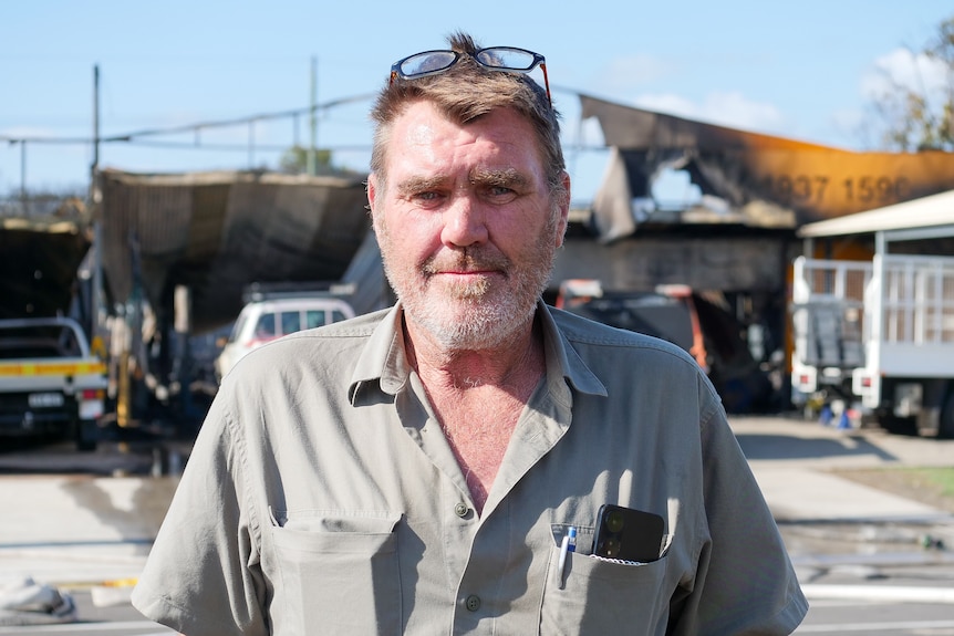 A close up of a man wearing a buttoned up shirt standing in front of a burnt tyre shop