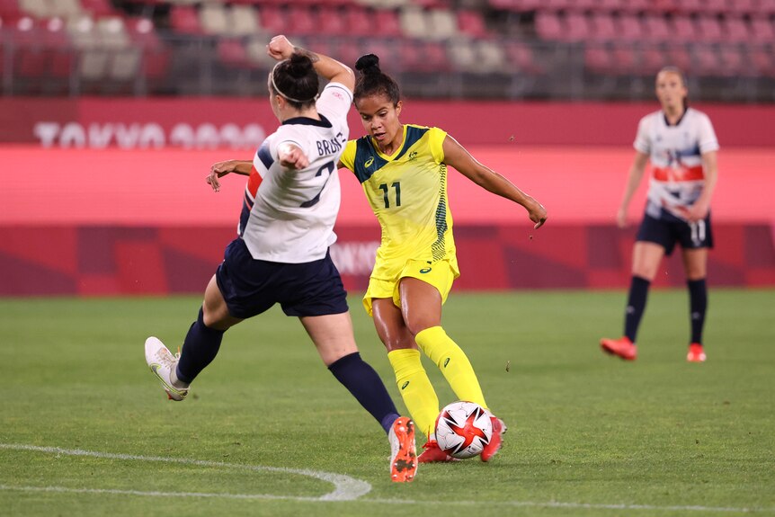 A woman wearing yellow kicks a ball in front of another woman wearing white