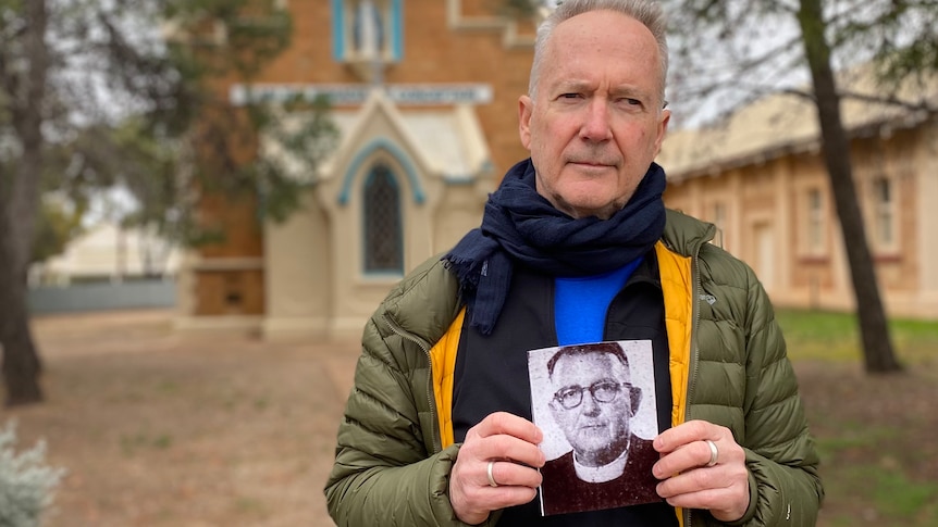 A man in a green puffer jacket stands outside a Catholic church holding an old photo of a priest