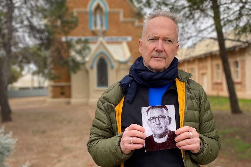 A man in a green puffer jacket stands outside a Catholic church holding an old photo of a priest