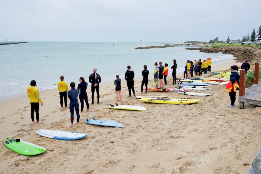 30 people in wetsuits stand alongside boards on the sand.