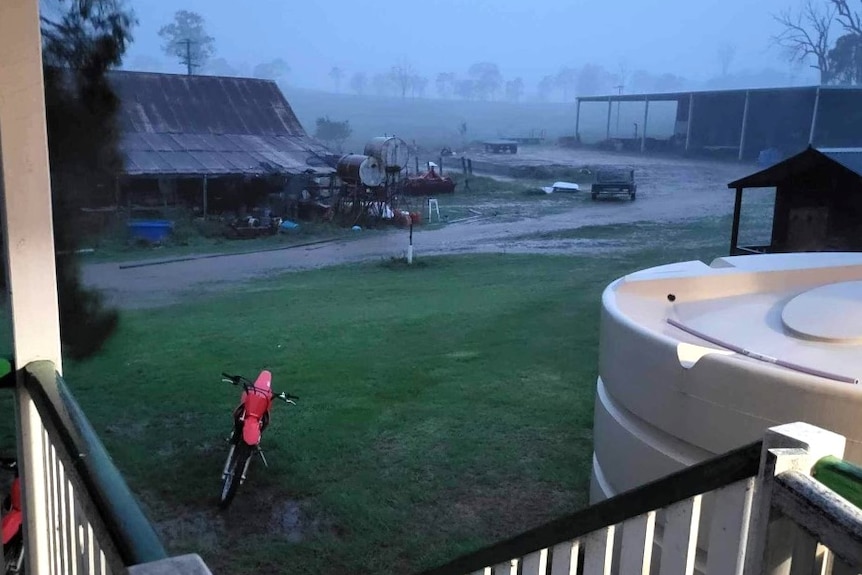 A rainy backyard with minor flooding on the driveway, a rain tank and motorbike, and sheds in the background.