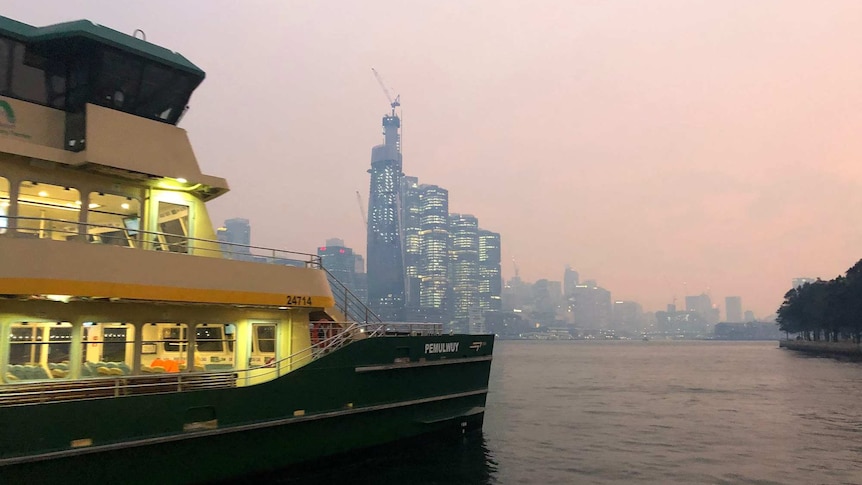 A ferry sets off from a wharf in front of a smoke city skyline.