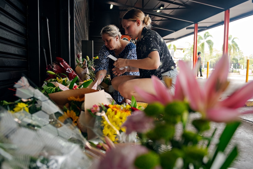 Two women kneeling down beside a long row of flowers in front of a bottle shop rollerdoor. 