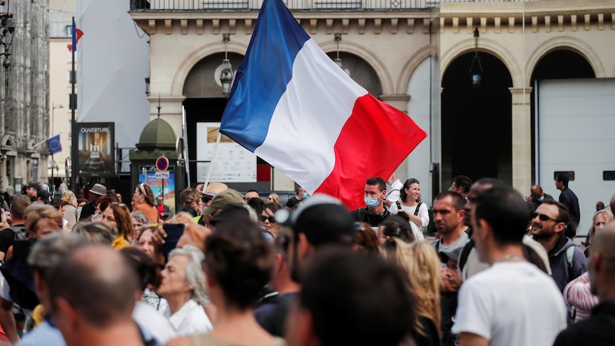 A French flag flies in the middle of a large crowd in a medieval square in a European city.