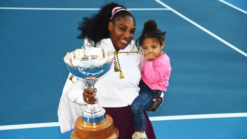 Serena Williams smiles while holding her daughter in one arm and a big silver trophy in the other