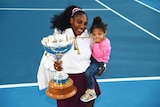 Serena Williams smiles while holding her daughter in one arm and a big silver trophy in the other