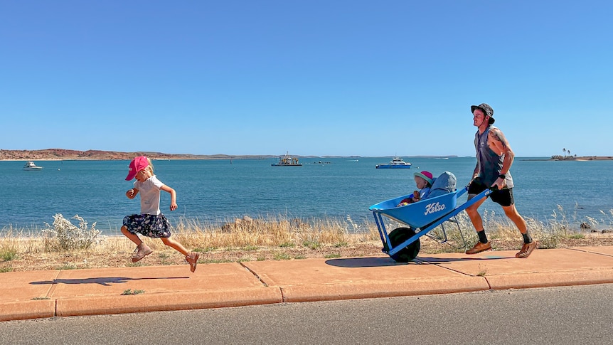 A young girl is running in front of her dad who is pushing her little sister in a blue wheelbarrow. They are next to the beach.