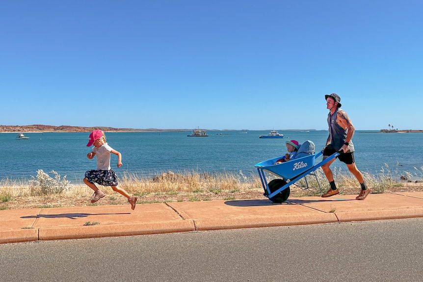 A young girl is running in front of her dad who is pushing her little sister in a blue wheelbarrow. They are next to the beach.