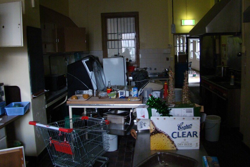 A room full of junk including a shopping trolley, old fridge, bottles, baskets, boxes and old cupboards