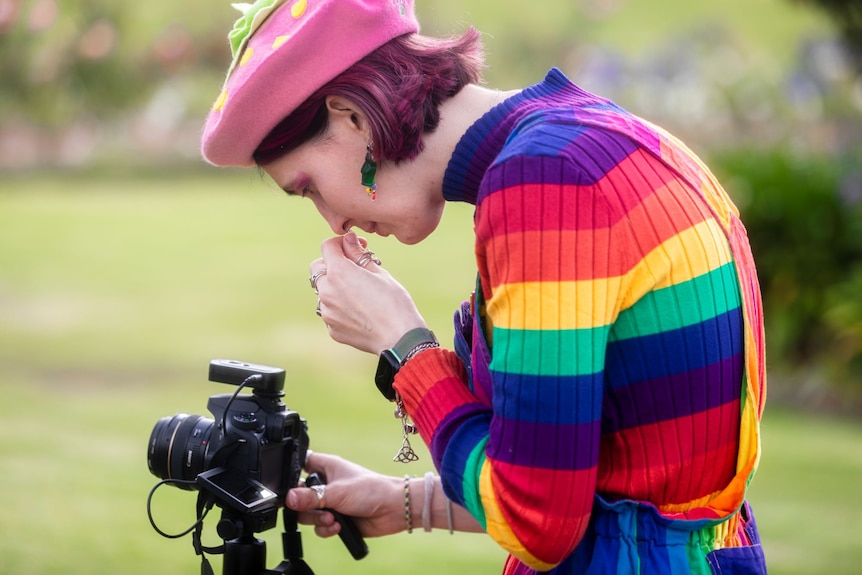 A woman in a pink beret, rainbow top and purple overalls prepares to take a photo with a camera.