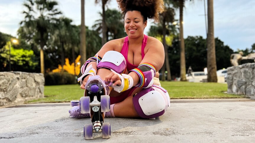 Yasmin laces up her purple skates while sitting on the ground in front of palm trees. She wears a pink activewear set.