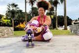 Yasmin laces up her purple skates while sitting on the ground in front of palm trees. She wears a pink activewear set.
