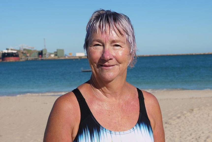 A close up shot of Julie smiling, she is in her bathers. The beach is on a sunny day is in the background.