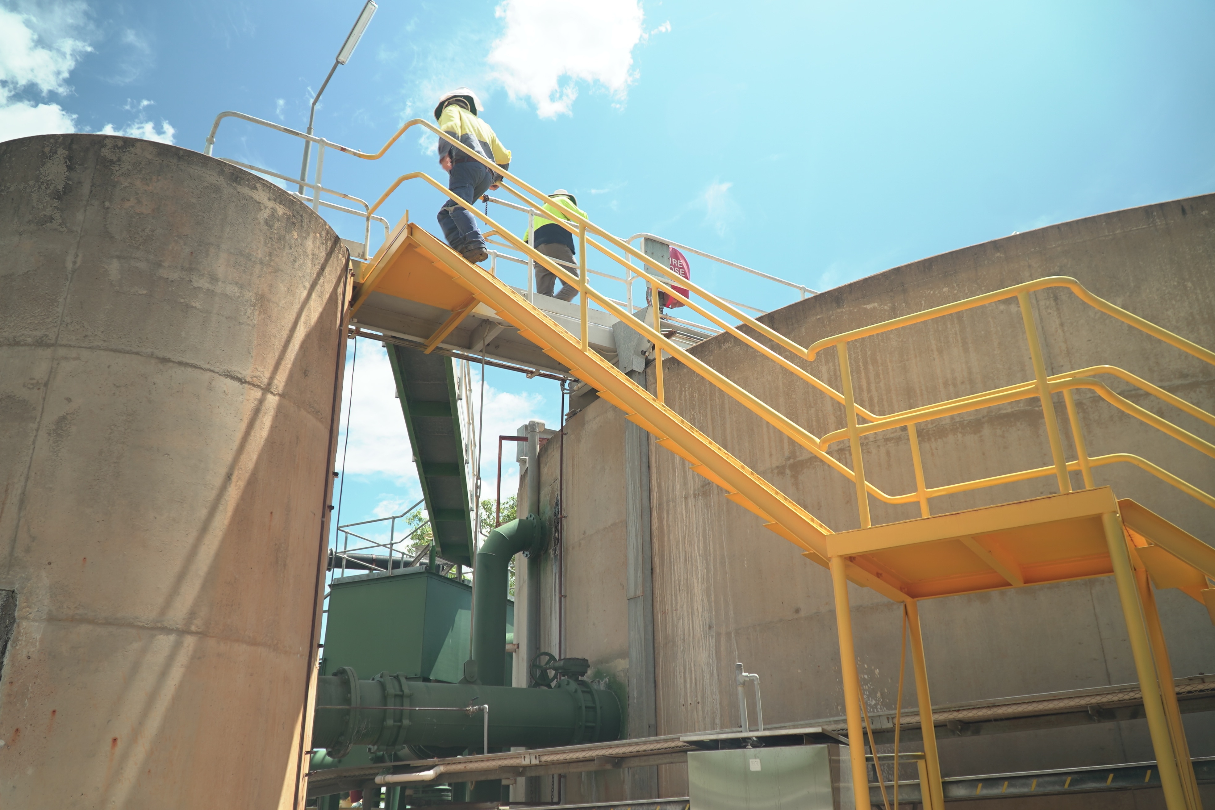 Dois trabalhadores em alta visibilidade subindo escadas entre dois tanques de concreto. 