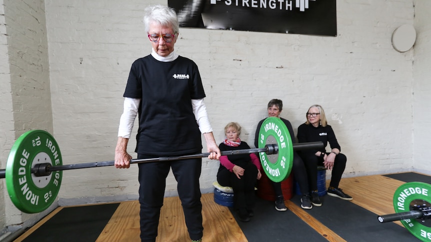 A older woman lifting a large weight with a group of fellow gym goers in the background
