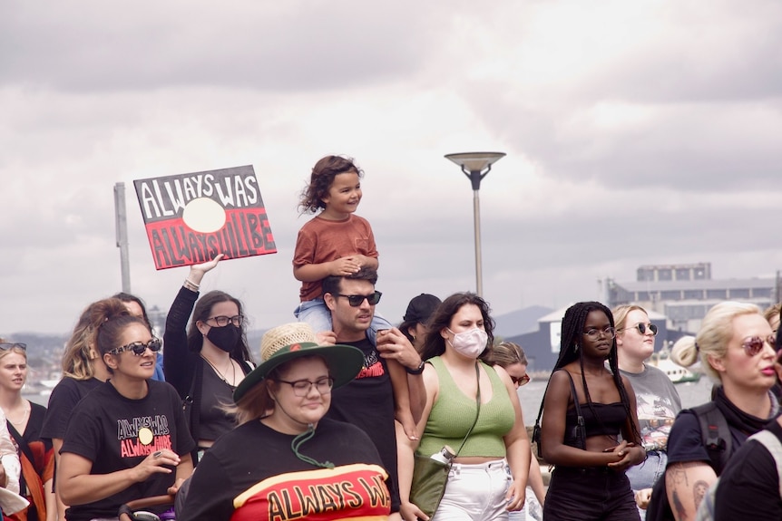 A young girl on a man's shoulders next to a sign that reads "Always was, always will be".