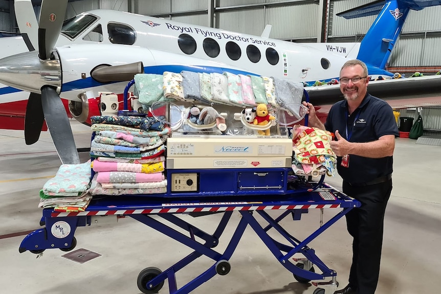 Man standing in front of a Royal Flying Doctor Service plane with trolley containing blankets and toys.  