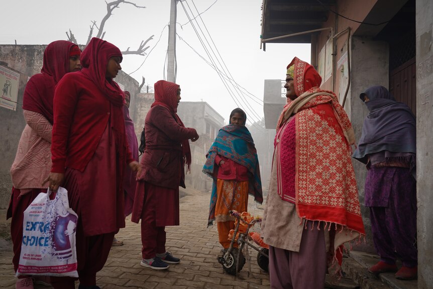 Women in maroon stand outside a doorway chatting with two villagers. One of the health workers is carrying a plastic bag