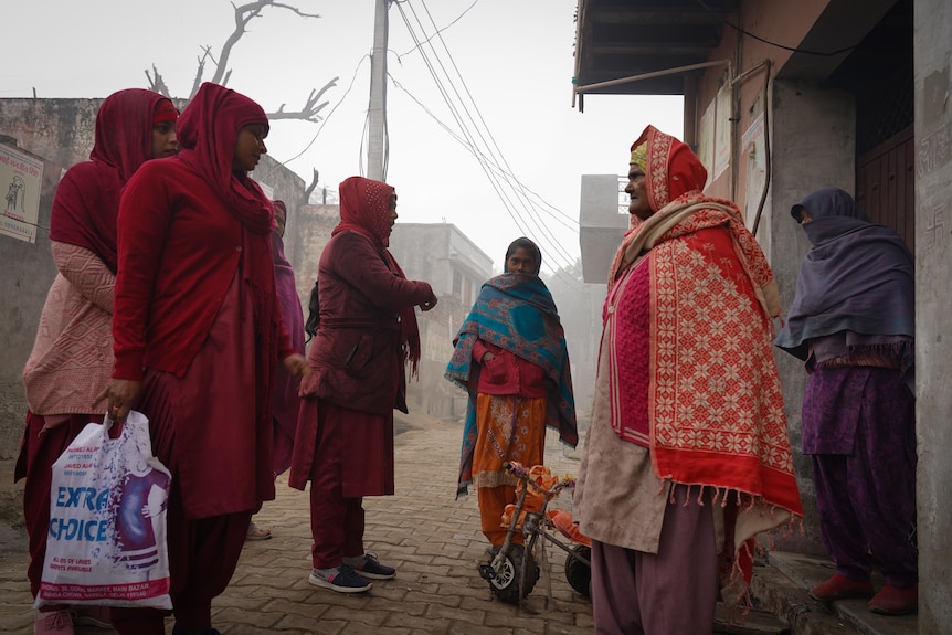 Women in maroon stand outside a doorway chatting with two villagers. One of the health workers is carrying a plastic bag