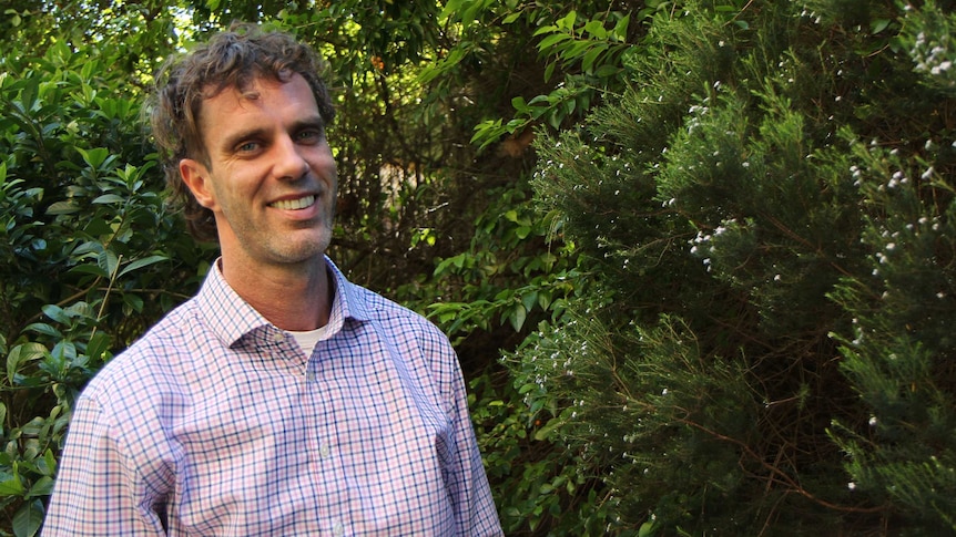 A man in a checked shirt stands in front of greenery.